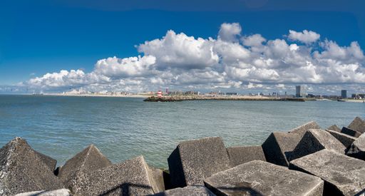 Large, geometric concrete blocks form a coastal barrier of Scheveningen harbor against the backdrop of the Hague skyline across the water.