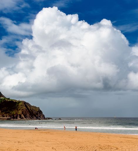A scenic beach with a dramatic cloud formation, people enjoying the water and sandy shore, and a rocky cliff in the background.