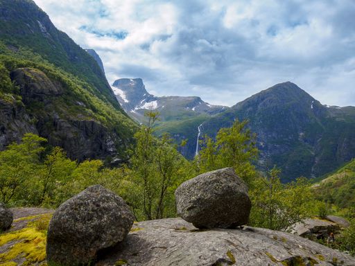 A scenic view of a lush green valley with rocky mountains and a waterfall in the distance under a cloudy sky.
