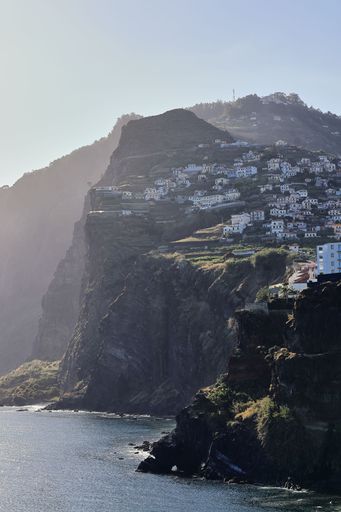 A scenic view of coastal cliffs with houses built on the hillside in Madeira, Portugal under a clear sky.