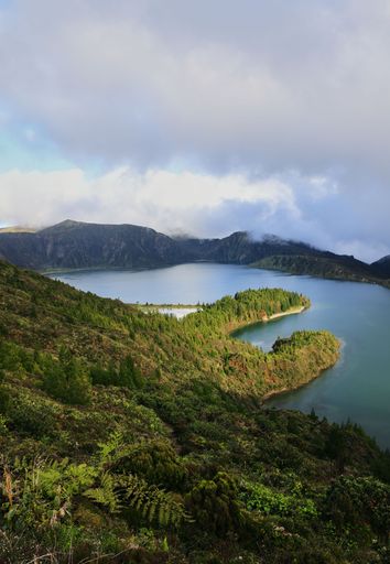 A scenic view of Lagoa do Fogo, a volcanic crater lake surrounded by lush greenery and mountains in the Azores, Portugal, under a cloudy sky.