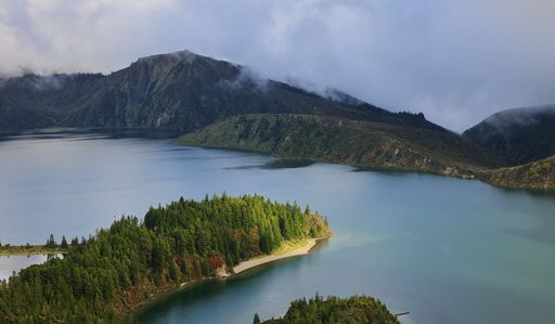 Scenic view of a lush green island in the middle of a calm lake surrounded by misty mountains under a cloudy sky.