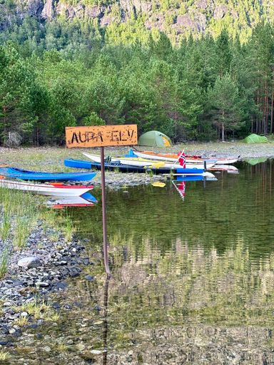 A serene riverside scene with kayaks, tents, and a sign reading 'Aura Elva' with a forested mountain backdrop.