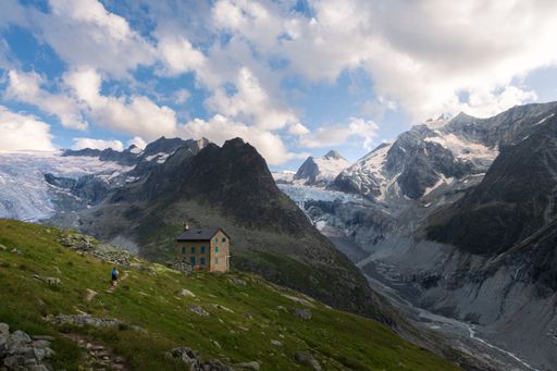 Summer landscape view of a hiker walking to a mountain hut as the sun is setting, surrounded by mountain peakss and glaciers and a cloudy sky