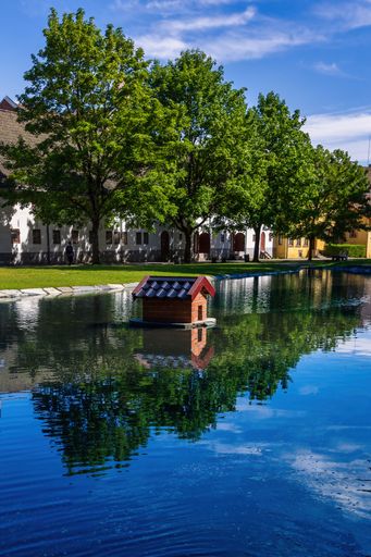 A serene pond with a small wooden house, surrounded by lush green trees and buildings reflecting in the water under a clear blue sky.
