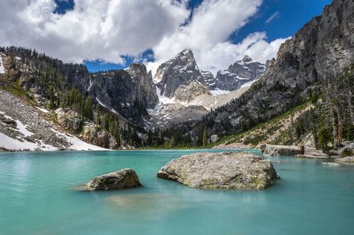Scenic view of a turquoise alpine lake surrounded by rugged mountains and pine trees under a partly cloudy sky.