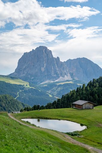 A serene mountain landscape featuring a small pond, a wooden cabin, and lush greenery under a partly cloudy sky in the Dolomites, Italy.