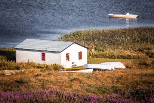 A serene lakeside scene featuring a small white house with red windows, surrounded by greenery and a boat in the water.