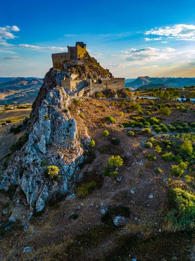 Aerial view of a historic castle on a rocky hilltop surrounded by scenic landscape under a clear blue sky.