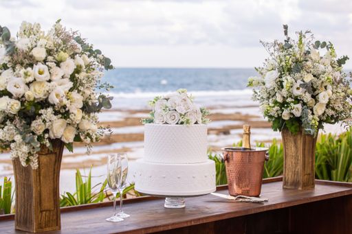 A beautiful wedding cake setup with floral decorations by the ocean, featuring champagne and glasses.