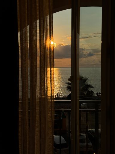 A beautiful sunset view through a window with sheer curtains, overlooking the sea and beach with silhouettes of people.