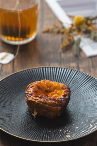 A close-up of a traditional custard egg tart on a black plate with a cup of tea in the background