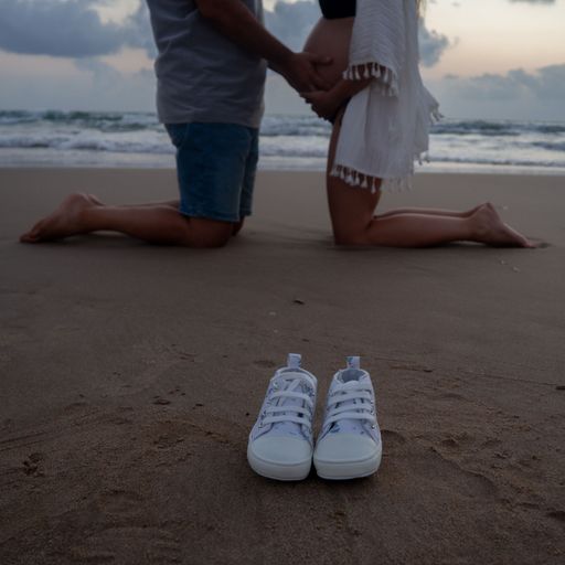 couple on their knees on the beach with their future baby's shoes in front of themERA