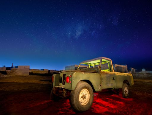 An old, rustic truck illuminated under a starry night sky in a desert village.