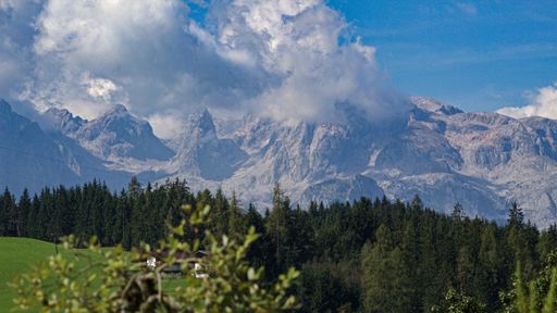 The Alpine Mountain range in Austria covered in snow and white clouds, with a green mountain in the foreground on a sunny day