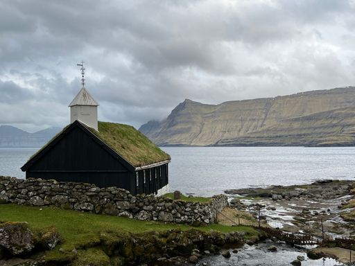A picturesque church with a grass roof by the sea, surrounded by mountains under a cloudy sky in the Faroe Islands.