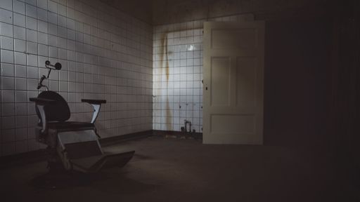 A dimly lit, abandoned room with white, tiled walls and a solitary dental chair in the center