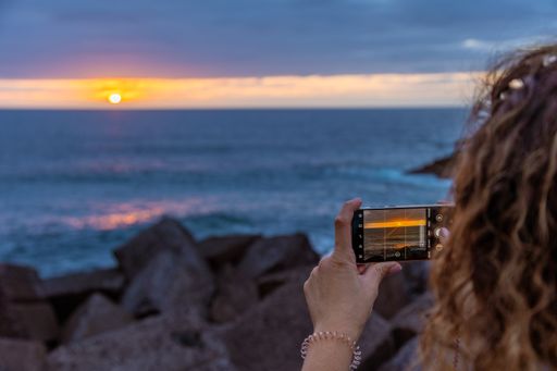 A curly woman photographing the sunset sky over the sea in summer