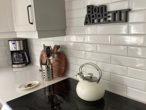 Modern kitchen interior with a coffee maker, utensils, and a kettle on a white countertop with a 'Bon Appetit' sign on the tiled backsplash.