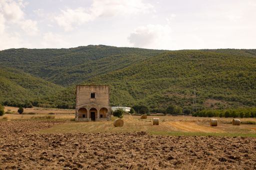 A rustic abandoned farmhouse in a plowed field with hay bales and green hills in the background under a partly cloudy sky.