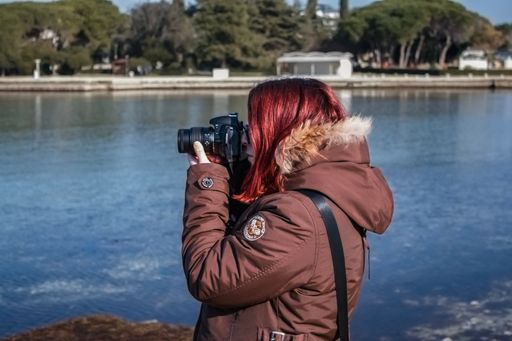 A young woman with red hair taking a photo by the water on a sunny day, wearing a brown winter jacket with a fur-lined hood.