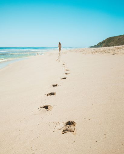 A woman walking along a pristine sandy beach leaving footprints behind her, with a blue sky in the background