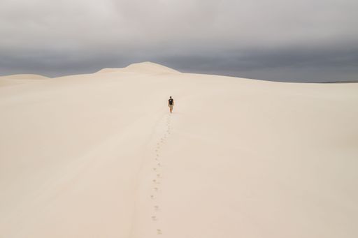 A solitary man walking on a vast sand dune under an overcast sky, leaving footprints behind.