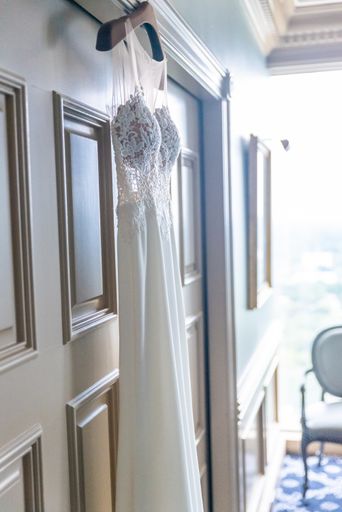 A vertical closeup shot of a bride's white dress hanging on a white door from a wooden hanger