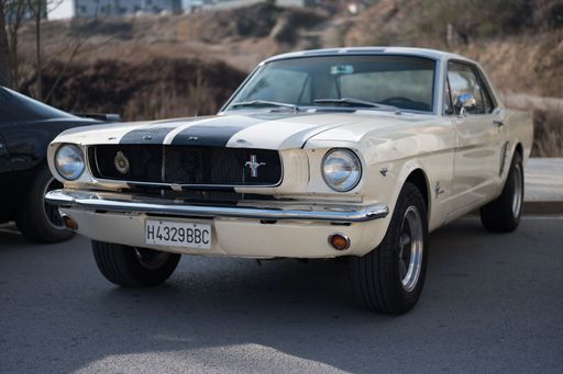 Front view of a classic first-generation Ford Mustang American sports car with a 289 cubic inch V8 engine. It is white with two blue stripes.