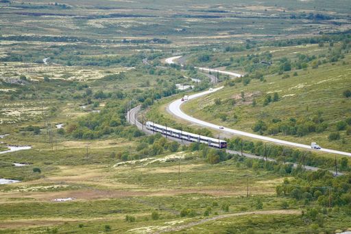 A scenic view of a train traveling through a lush green landscape with winding roads and sparse vehicles.