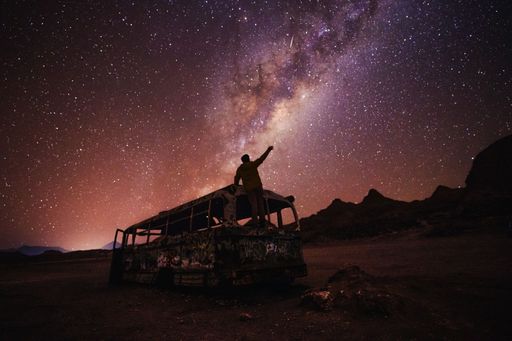 A person standing on an abandoned bus under a starry night sky with the Milky Way visible, creating a stunning and atmospheric scene.