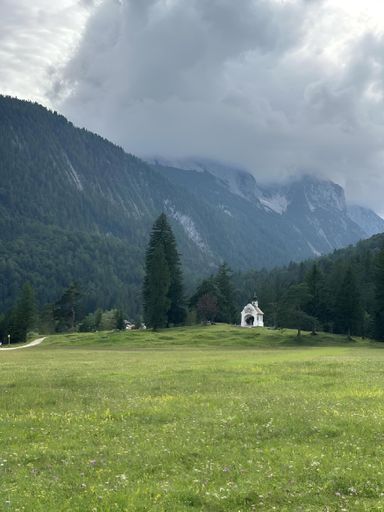 A serene landscape featuring a small chapel amidst lush green fields and towering mountains under a cloudy sky.