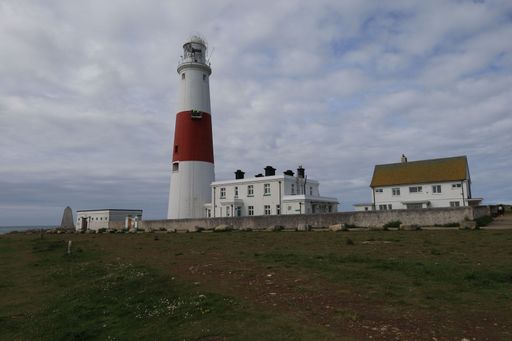 A tall, white lighthouse with a red band stands on a cliff overlooking the ocean. A white house and a smaller building are located nearby.