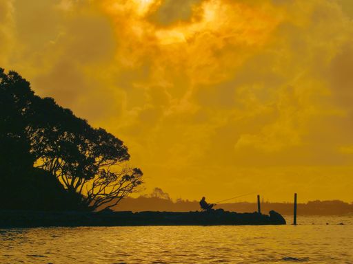 A silhouetted fisherman fishing and relaxing in the evening on a chair beside the estuary waters at sunset with trees behind him