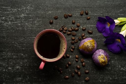 A top view of a cup of coffee, roasted coffee beans, sweets, and flowers on the grunge dark surface