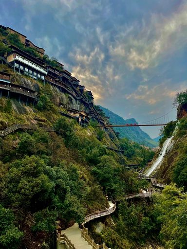 A scenic view of traditional houses built on a mountain slope with a suspension bridge and lush greenery in the foreground.