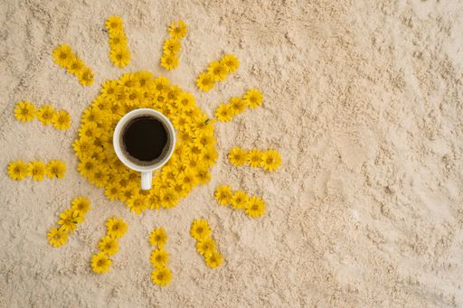 A top view of a cup of coffee surrounded by yellow flowers forming a sun on a rock surface
