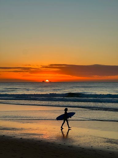 A silhouette of a surfer walking on the beach with a surfboard during a vibrant sunset.