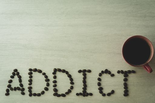 A top view of roasted beans formed as addict text beside a cup of coffee with copy space