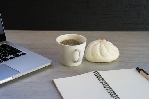 A cup of hot coffee, steamed bun, notepad and a laptop on a desk during the break