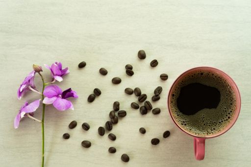 A top view of a cup of coffee with purple flowers and roasted beans  on the surface