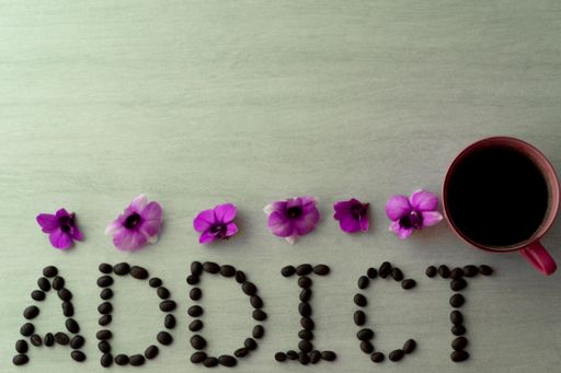 A top view of roasted beans formed as addict text with purple flowers beside a cup of coffee