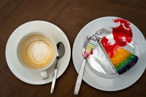 A top view of a delicious slice of rainbow-colored cake next to a cup of cappuccino coffee on a wooden table