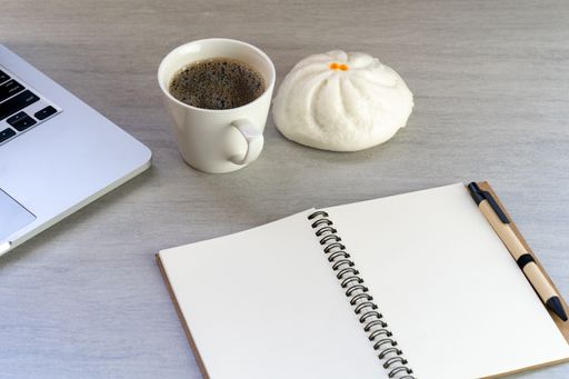 A top view of a hot bun and a cup of coffee beside a laptop and an open notebook with copy space