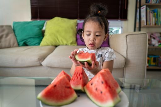 A selective focus shot of a Southeast Asian girl eating biting from big watermelon slices indoors