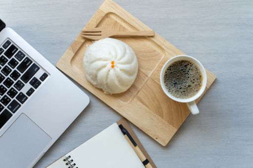 A top view of a hot bun and a cup of coffee on a wooden board beside a laptop