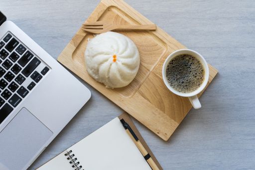 A top view of a coffee with a dessert next to the laptop and a notebook on an office table