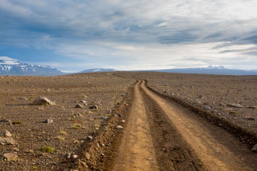 A remote dirt road winding through a barren landscape with distant snow-capped mountains under a cloudy sky.