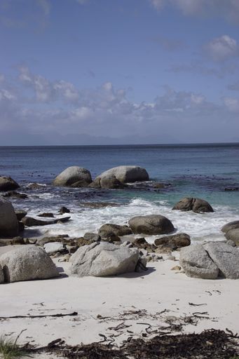 A serene beach scene with large rocks on the shore, clear water, and a blue sky with some clouds.