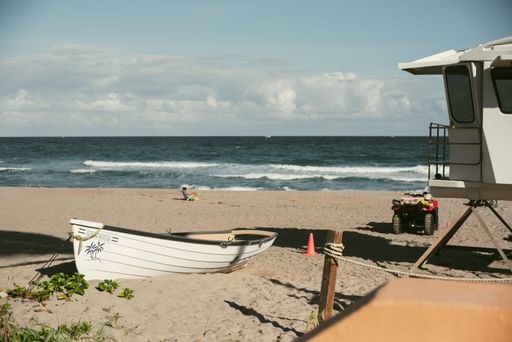 A wooden boat next to cabin on the sandy shore under a clear sky and a person sitting in front of the sea in the background
